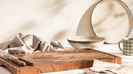 A wooden cutting board next to ceramic plates and silverware wrapped in a linen napkin, bathed in sunlight.