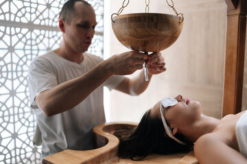 Portrait of spa mater gently guiding oil stream on female client forehead during extraordinary Shirodhara treatment, Ayurvedic healing technique in wellness salon. Concept of body care, rehabilitation