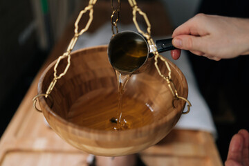 Close-up cropped shot of unrecognizable spa specialist pouring hot aromatic oil into bowl during preparation for exotic Shirodhara treatment, Ayurvedic healing technique in wellness center.