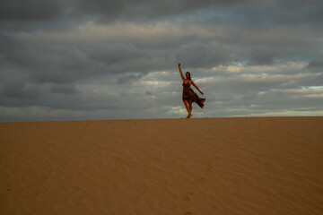 A woman is standing on a sandy hill, wearing a red dress and waving her arms. The sky is cloudy,...