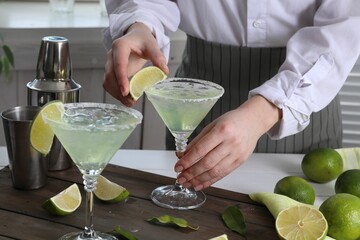 Bartender putting piece of lime onto glass with delicious Margarita cocktail at wooden table,...