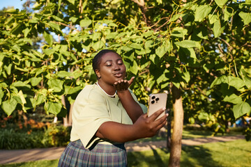 Naklejka na ściany i meble Plus size African American lady stands under a tree, holding a cell phone on a sunny day.