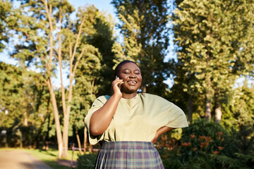 Naklejka na ściany i meble Plus size African American woman in casual attire, embracing body positivity, chats on a cell phone in a park on a sunny day.
