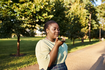 Plus-size African American woman stands on roadside, chatting on phone in summer heat, embracing body positivity.