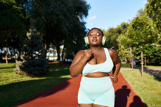 A curvy African American woman in sportswear running and wearing headphones, enjoying her outdoor exercise routine.