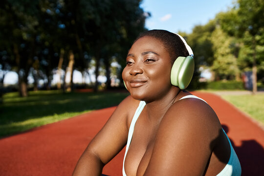 An African American woman in sportswear sits, wearing headphones, immersed in music, embodying body positivity while outdoors. - Powered by Adobe