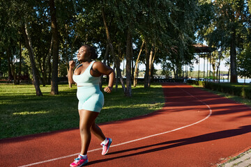 Obraz premium An African American woman in sportswear energetically runs along a track in a lush park setting, embodying positivity and confidence.