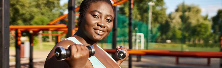 An African American woman in sportswear exercises with dumbbells in a lush park setting.
