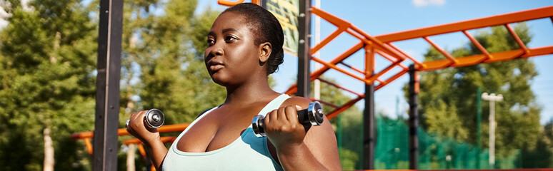 An African American woman in sportswear joyfully lifts dumbbells in front of a colorful playground.