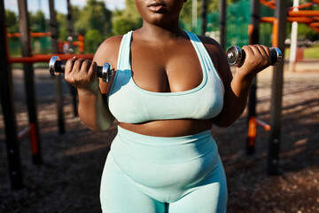 A body-positive African American woman in a blue sports suit confidently lifts two dumbbells outdoors.