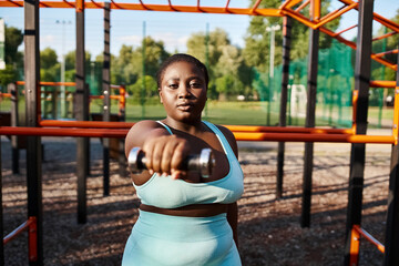 A curvy African American woman in a blue sportswear confidently holds a metal dumbbell in her hands, exuding grace and strength.