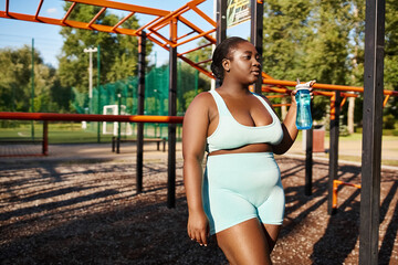 An African American woman in sportswear stands in a park, holding a water bottle, taking a refreshing break from her outdoor workout.