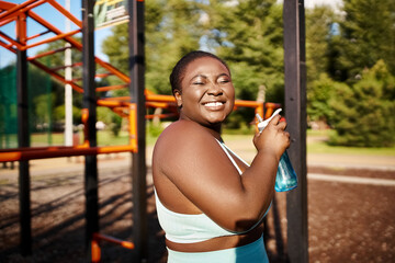 African American woman in sportswear stands at playground holding bottle of water.