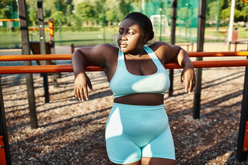 An African American woman in sportswear happily stands in front of a playground, embodying body positivity and empowerment.