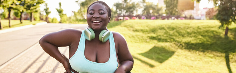 A stylish African American woman with a curvy figure, wearing a blue top and headphones, enjoying music outdoors.