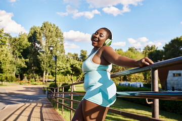 An African American woman with curly hair, wearing a blue and white dress, leans elegantly on a rail while enjoying some music.