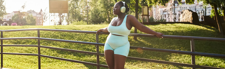 African American woman in a blue outfit, leaning gracefully on a fence, enjoying music through headphones in an outdoor setting.