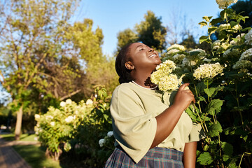 A plus size African American woman in casual attire stands gracefully next to a bush of vibrant flowers outdoors in the summer.