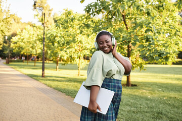 A plus-size African American woman in casual attire, wearing a green shirt and plaid skirt, outdoors in the summer.