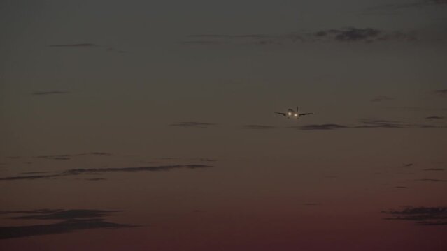 The plane is captured in flight with the landing gear released and the headlights, on against the background of a twilight sky with layered clouds and a gradient of sunset colors from orange to blue