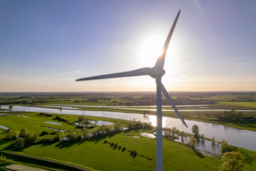 Blades of wind turbine slowly rotating with sun directly behind creating halo and flares in Dutch river IJssel landscape. Aerial circular economy industry and sustainability concept in The Netherlands