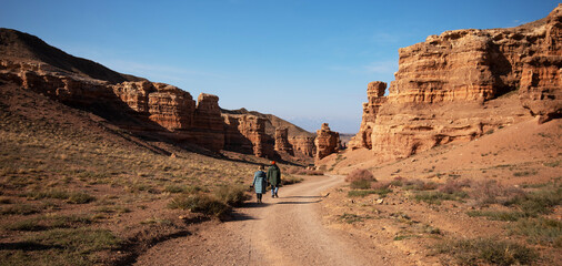 two individuals are walking along a dirt path in a desert canyon. The canyon walls, towering on...