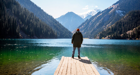 A man stands on a wooden pier looking out at a tranquil mountain lake, surrounded by forested slopes and snow-capped peaks under a clear blue sky. - obrazy, fototapety, plakaty