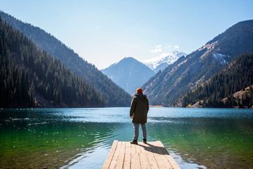 A man stands on a wooden pier looking out at a tranquil mountain lake, surrounded by forested slopes and snow-capped peaks under a clear blue sky.