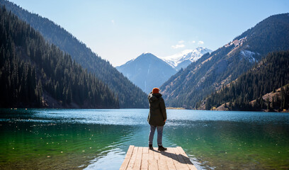 A man stands on a wooden pier looking out at a tranquil mountain lake, surrounded by forested slopes and snow-capped peaks under a clear blue sky.