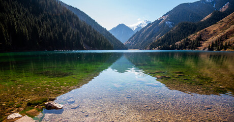 Crystal-clear waters of a mountain lake reveal stones beneath, leading to forested hills and...