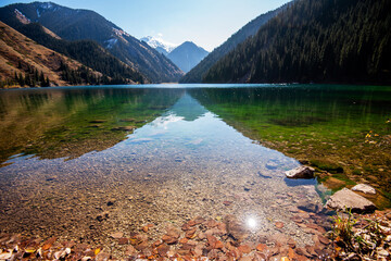 Crystal-clear waters of a mountain lake reveal stones beneath, leading to forested hills and...