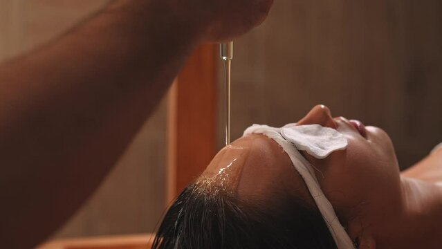 Closeup of pouring oil on female forehead during exotic Shirodhara treatment, Ayurvedic healing technique . Serene young woman at massage session with aromatic oil dripping on forehead and hair.