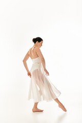 Fototapeta na wymiar A young woman in a flowing white dress gracefully dances in a studio setting against a white background.