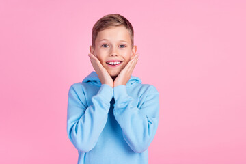 Photo of astonished cheerful boy toothy smile hands touch cheeks empty space isolated on pink color...