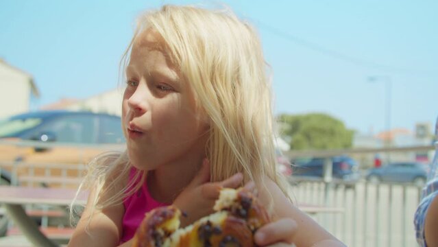 A blonde girl in a pink shirt eating a bun with raisins on the street against the background of a blurred city