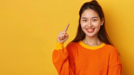 Beautiful Asian woman in orange smiles and points with her hand, comparing two options and showing an empty space in a yellow studio.