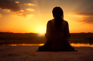 Prayer concept. Silhouette of a pretty African american woman in praying pose. Set against a vibrant sunset sunrise sky. Clasped hands. Also related to vicarious, word of god, zion, reverence, divine