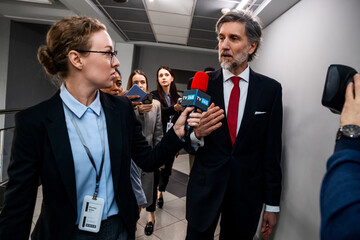 Mature organization representative answers press questions and gives interview for TV news walking in parliament corridor. Party representative surrounded by crowd of TV journalists. Press campaign.