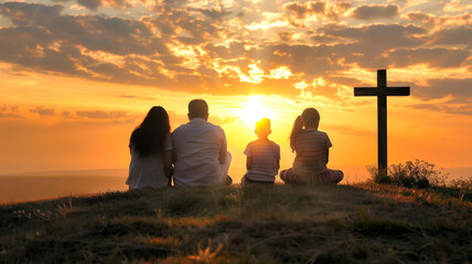 A religious family of four sits on a hillside looking at the sun as it sets with a cross next to them