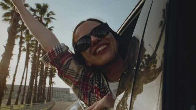 Excited cheerful girl smiling. Female is enjoying a vacation in a tropical city by the sea or ocean. Woman in sunglasses, having fun on exotic island seaside road. Palm trees are reflected in the car