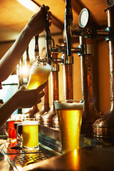 Close-up of bartender's hands in action, pouring frothy lager beer from the taps into glass. Pub...