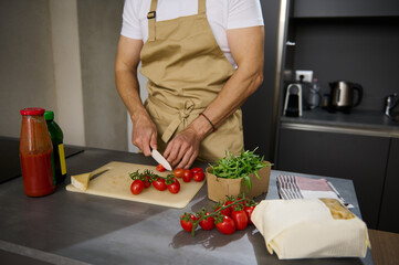 Close-up view of a male chef in beige apron, chopping tomato cherry on a cutting board. Ingredients on the kitchen table. Man cooking healthy salad in the minimalist home kitchen interior