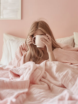 A girl in bed under a pink blanket with a coffee mug covering her face. Cozy morning retreat with coffee comfort. Minimal pastel pink combination.