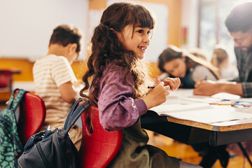 Education and child development. Little girl smiles while sitting in a class with her teacher and fellow students - obrazy, fototapety, plakaty