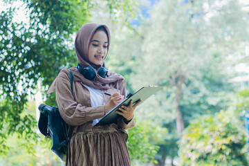Young asian girl in hijab smiling writing on book and carrying bag clipboard standing in outdoor park. female students for the themes of education, technology and advertising