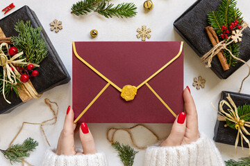 Hands with red envelope near gift boxes, fir branches and Christmas decor, Mockup