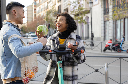 An eco-friendly couple laughing and making jokes on the street. She rides an electric scooter and he recycles bags. - Powered by Adobe