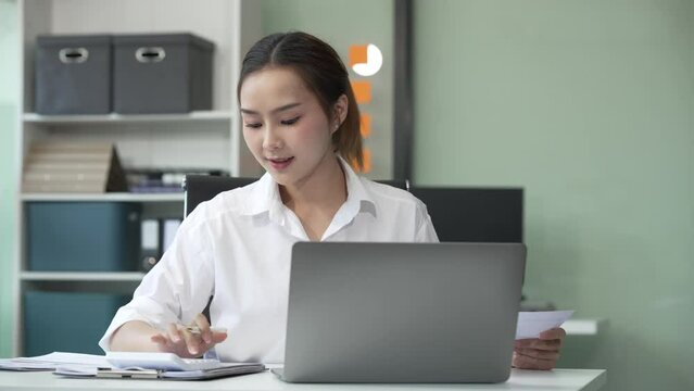 Asian young businesswoman, a Financial Advisor, works happily in her office. She uses her laptop and mobile phone to analyze charts and communicate financial strategies. Red formal suit