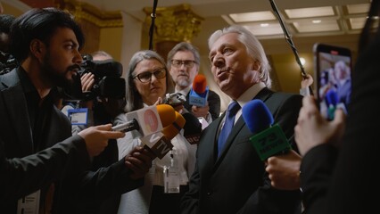 Politician answers press questions and gives interview on television political program. Senior diplomat surrounded by crowd of journalists in government building. Press conference. Hot button issues. - obrazy, fototapety, plakaty