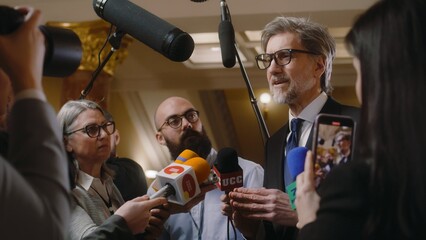 Politician answers press questions and gives interview for television news. Mature diplomat surrounded by crowd of journalists in parliament hall. Press conference. Inspirational speech. Slow motion. - obrazy, fototapety, plakaty